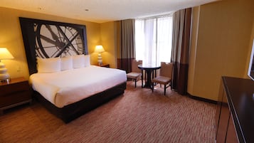 Habitación de hotel con una cama grande, dos lámparas de noche, una mesita con una silla y un amplio ventanal con cortinas.