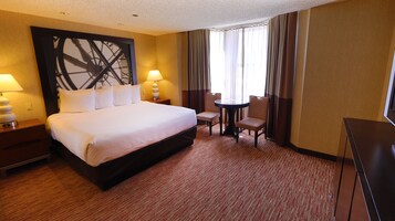 Habitación de hotel con una cama grande, dos lámparas de noche, una mesita con una silla y un amplio ventanal con cortinas.
