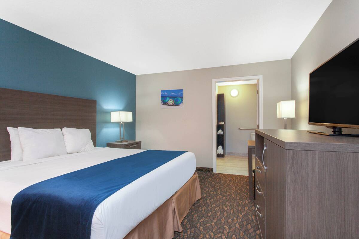 A hotel room with a large bed, a desk with a television, a nightstand with a lamp, and a door leading to another room.