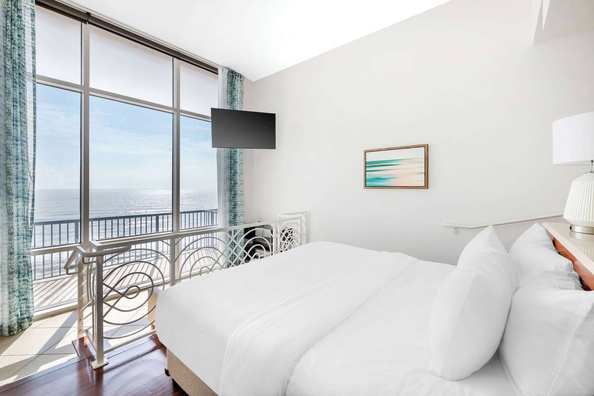 A hotel room with a large window, a bed, a TV, and a bedside lamp.
