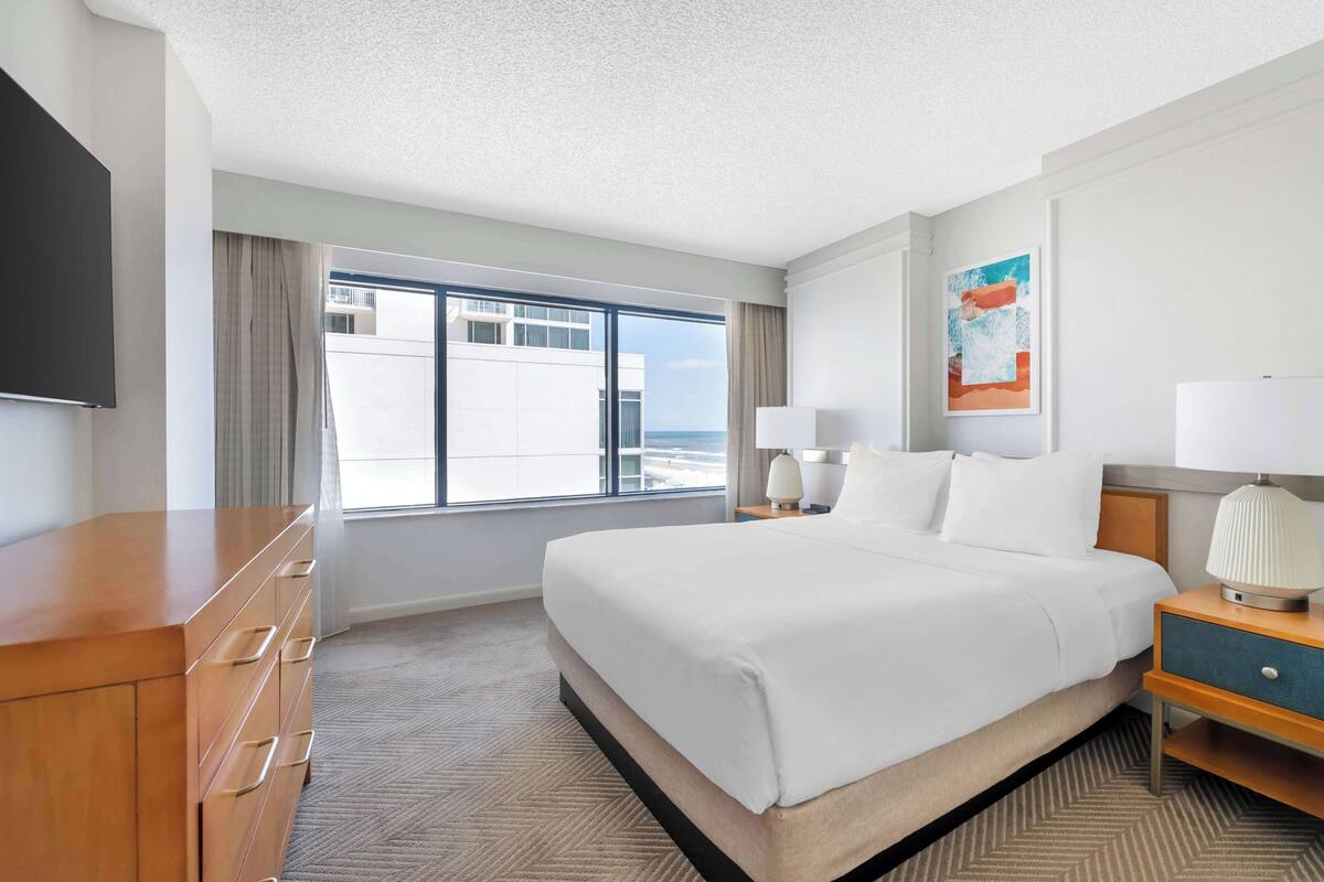 A hotel room with a large bed, a wooden dresser, a flat-screen TV, a window with curtains, and a painting on the wall.