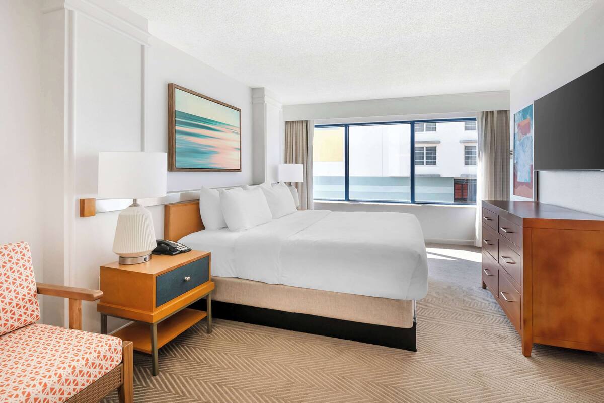 A hotel room with a large bed, a desk with a lamp, a chair, a dresser, and a television.
