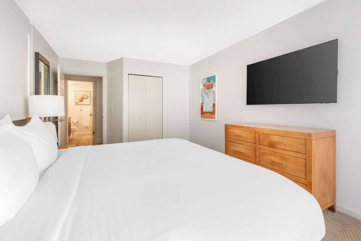 A hotel room with a large bed, a wooden dresser, a flat-screen TV mounted on the wall, and a door leading to a bathroom.
