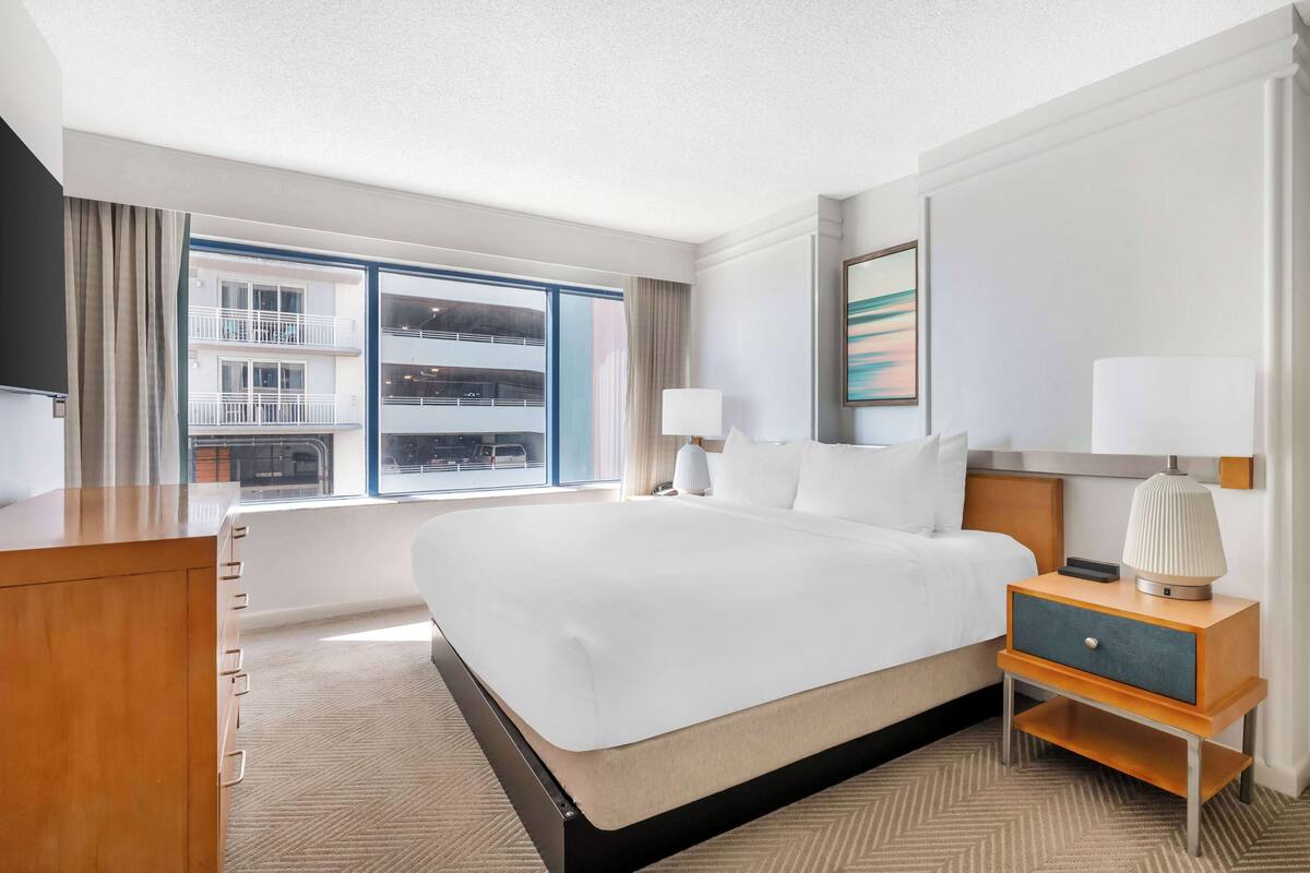 A hotel room with a large bed, a wooden nightstand, a TV, and a window with a view of a building.