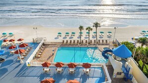 Un espace piscine doté de chaises longues et de parasols, avec vue sur une plage bordée de palmiers.