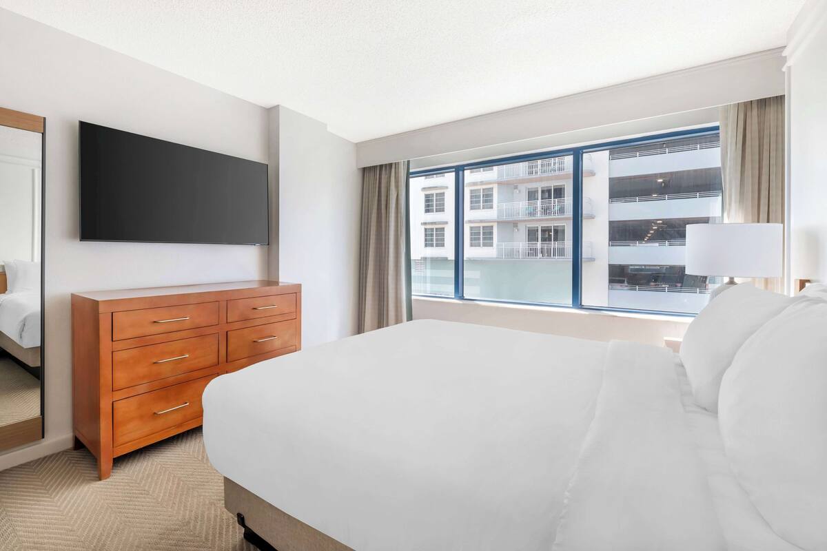 A hotel room with a large bed, a wooden dresser, a flat-screen TV, and a large window with a view of a building.