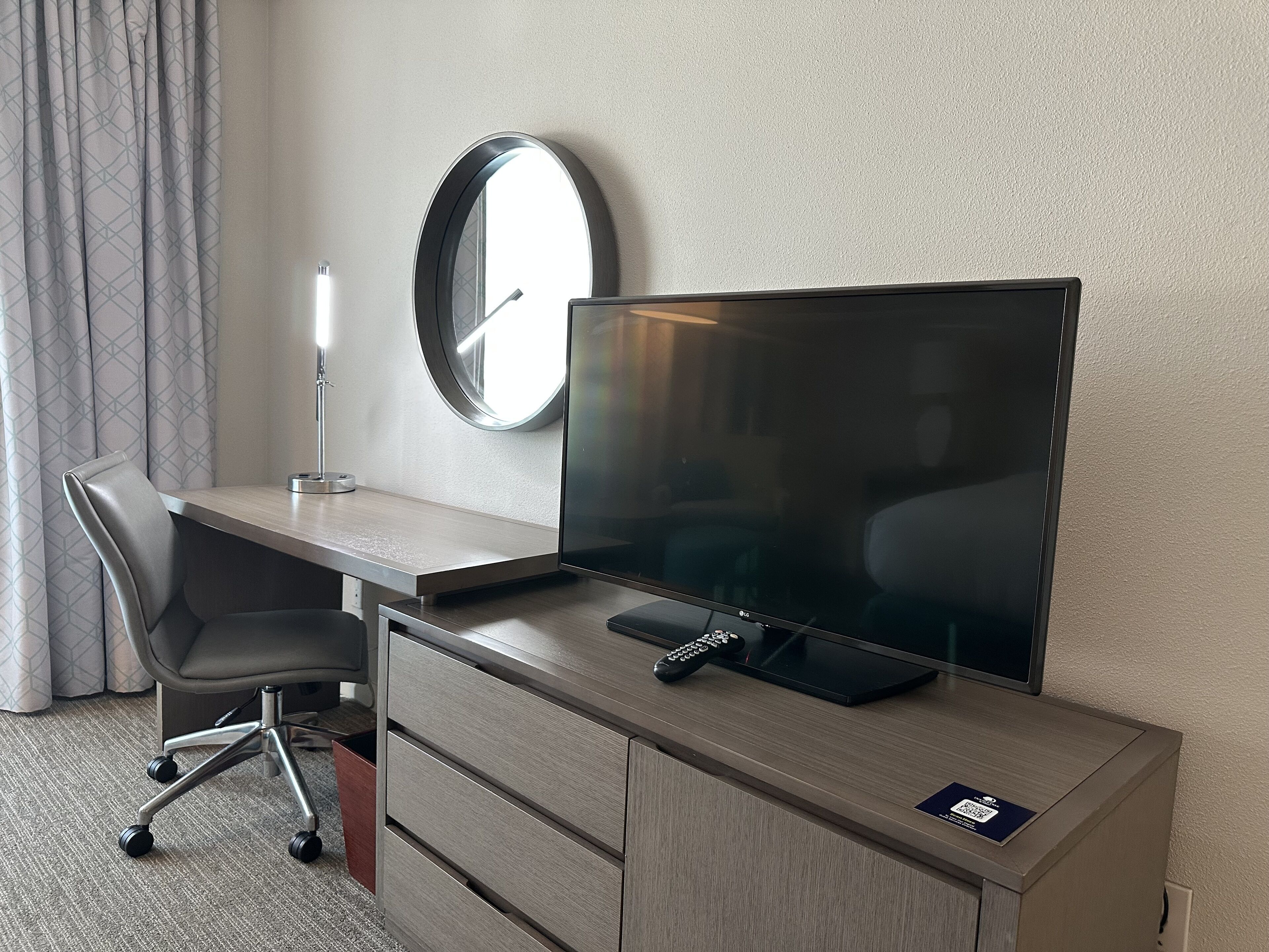 Ein Hotelzimmer mit einem Holztisch, einem modernen Sessel, einem Flachbildfernseher, einer Fernbedienung und einem Wandspiegel.