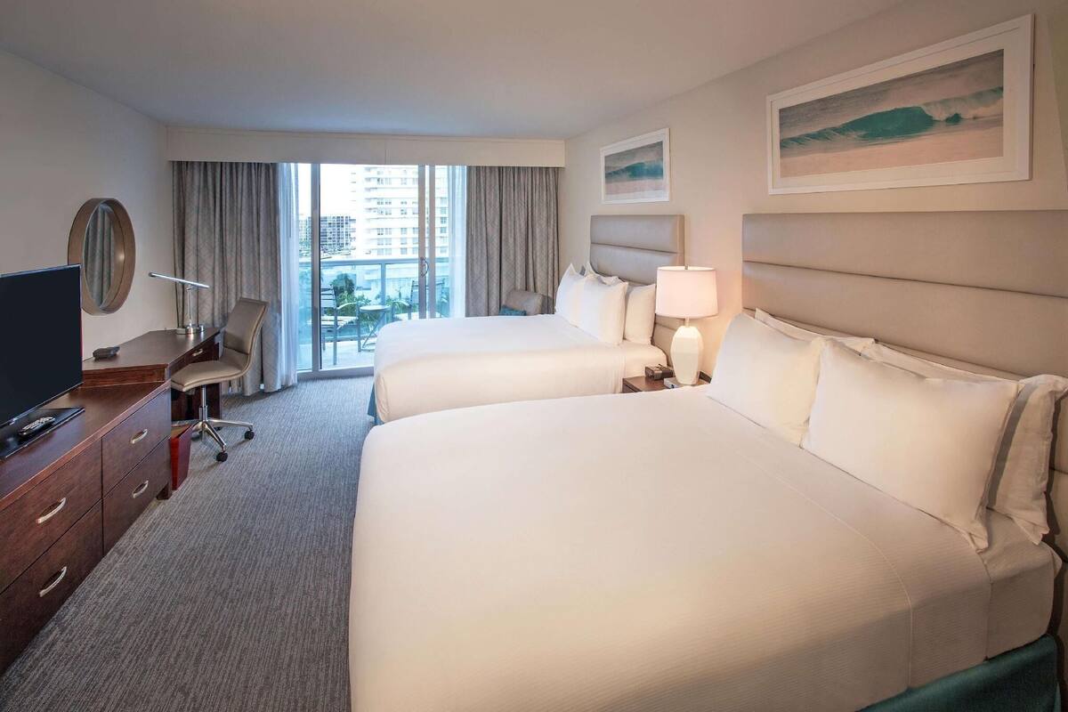 Ein Hotelzimmer mit zwei Betten, einem Schreibtisch mit Fernseher, einem Sessel, einer Schreibtischlampe und einem Fenster mit Blick auf Gebäude.