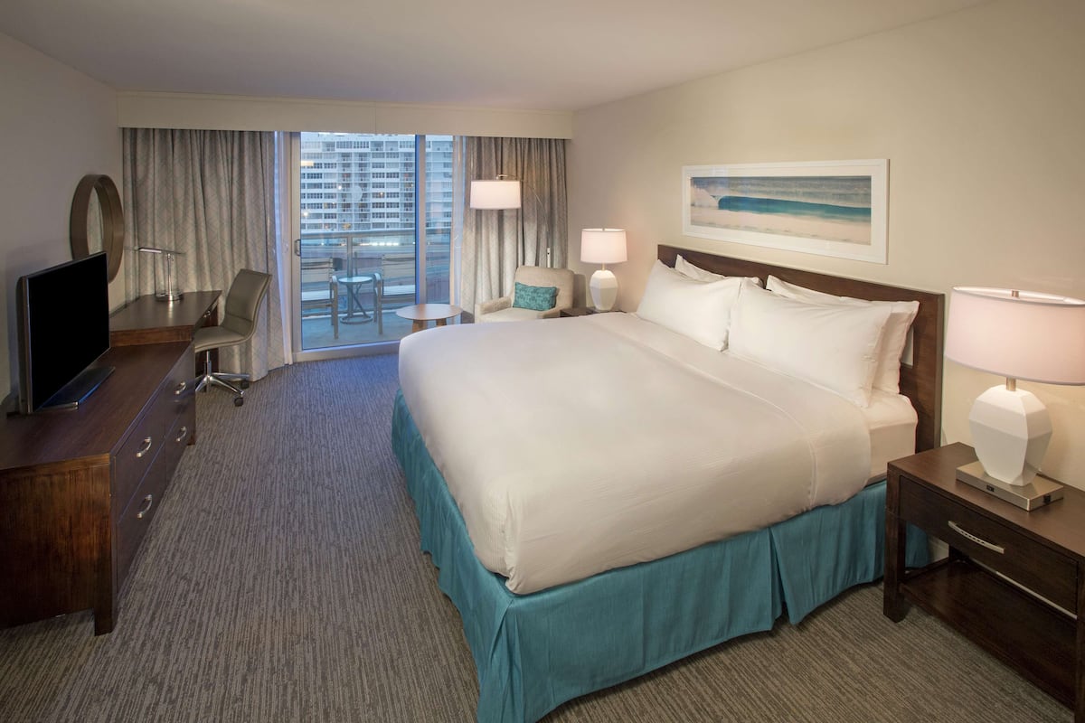 Ein Hotelzimmer mit einem großen Bett, einem Schreibtisch mit Fernseher, einem Sessel, einem kleinen Tisch und einem Balkon mit Blick auf die Stadt.