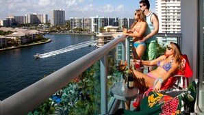 Un balcón con vistas a un río y al paisaje urbano, una mesa con bebidas y una persona recostada.