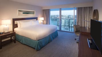 A hotel room with a large bed, a desk with a TV, and a view of a cityscape through large windows.