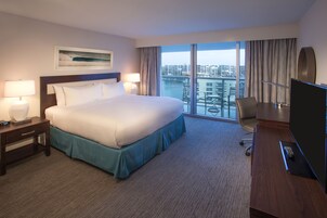 A hotel room with a large bed, a desk with a TV, and a view of a cityscape through large windows.