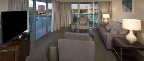 A modern hotel room with a large TV, comfortable seating, a small table, and a view of the cityscape.