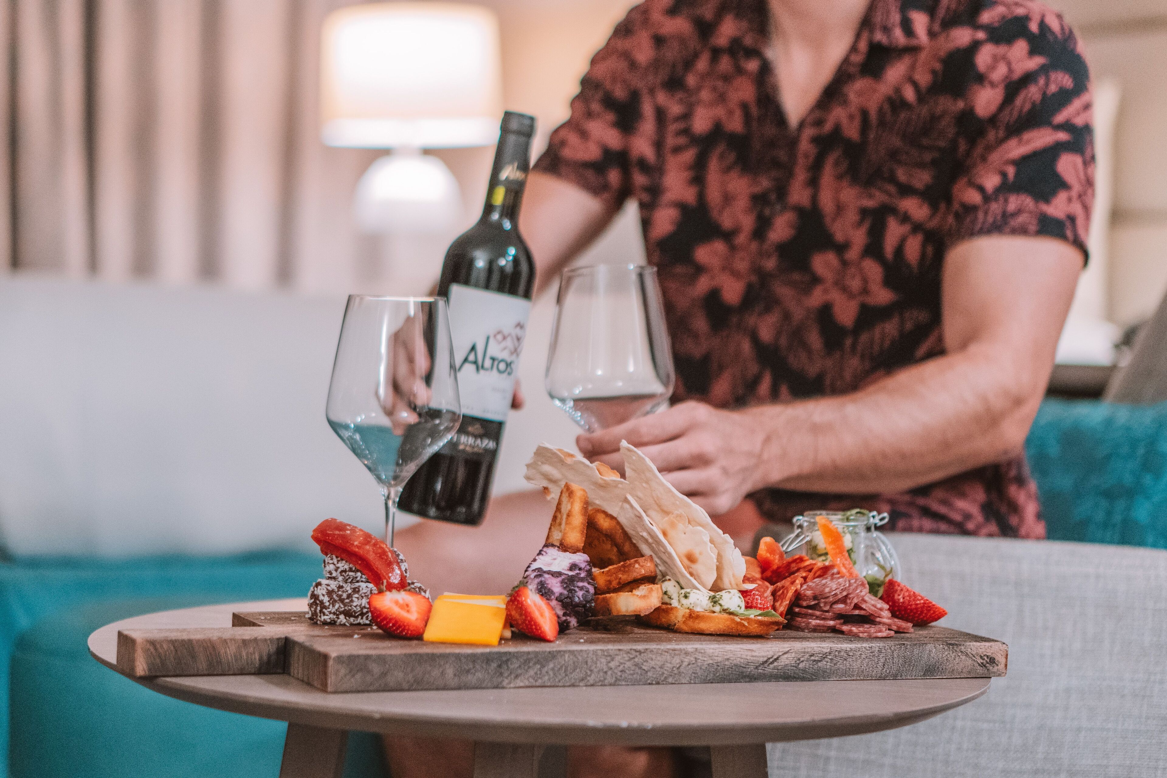Une personne verse du vin dans des verres sur une table où sont disposés divers fromages, fruits et pains.