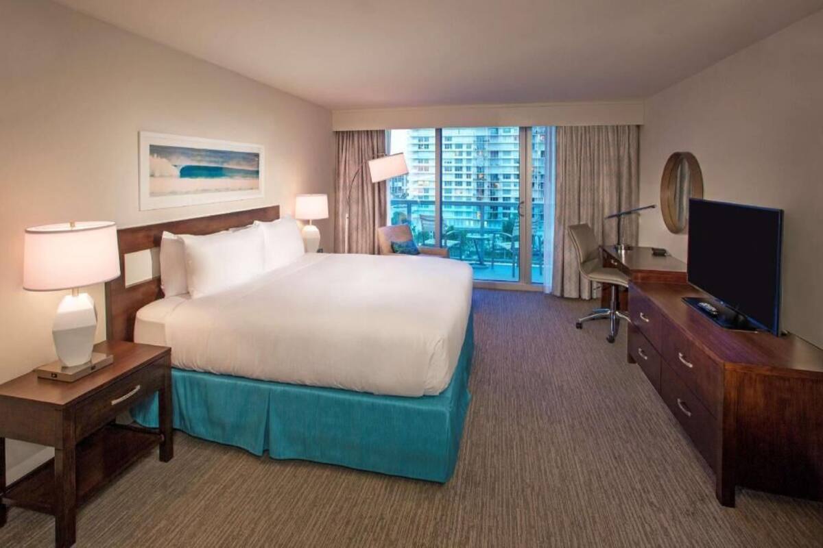 Ein Hotelzimmer mit einem großen Bett, einem Schreibtisch mit Fernseher, einem Sessel und Blick auf Gebäude durch das Fenster.