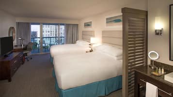 Habitación de hotel con una cama grande, un escritorio con computadora, una silla, televisor, baño con espejo y vistas a la ciudad.