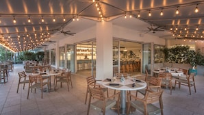 Un espacio de comedor al aire libre con mesas redondas y sillas de madera, ventiladores de techo e iluminacion con luces colgantes.