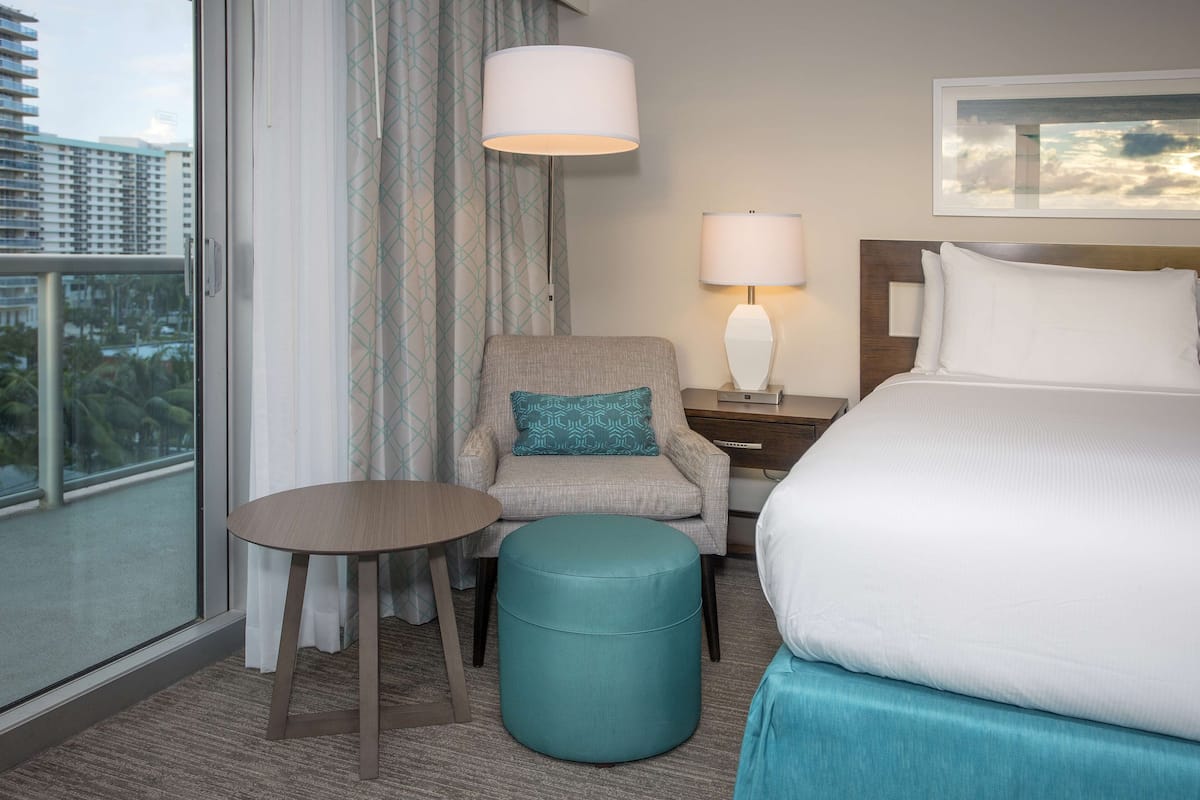 Ein Hotelzimmer mit einem Bett, einem Sessel, einem runden Tisch, einer Lampe und Blick auf Gebäude durch ein Fenster.