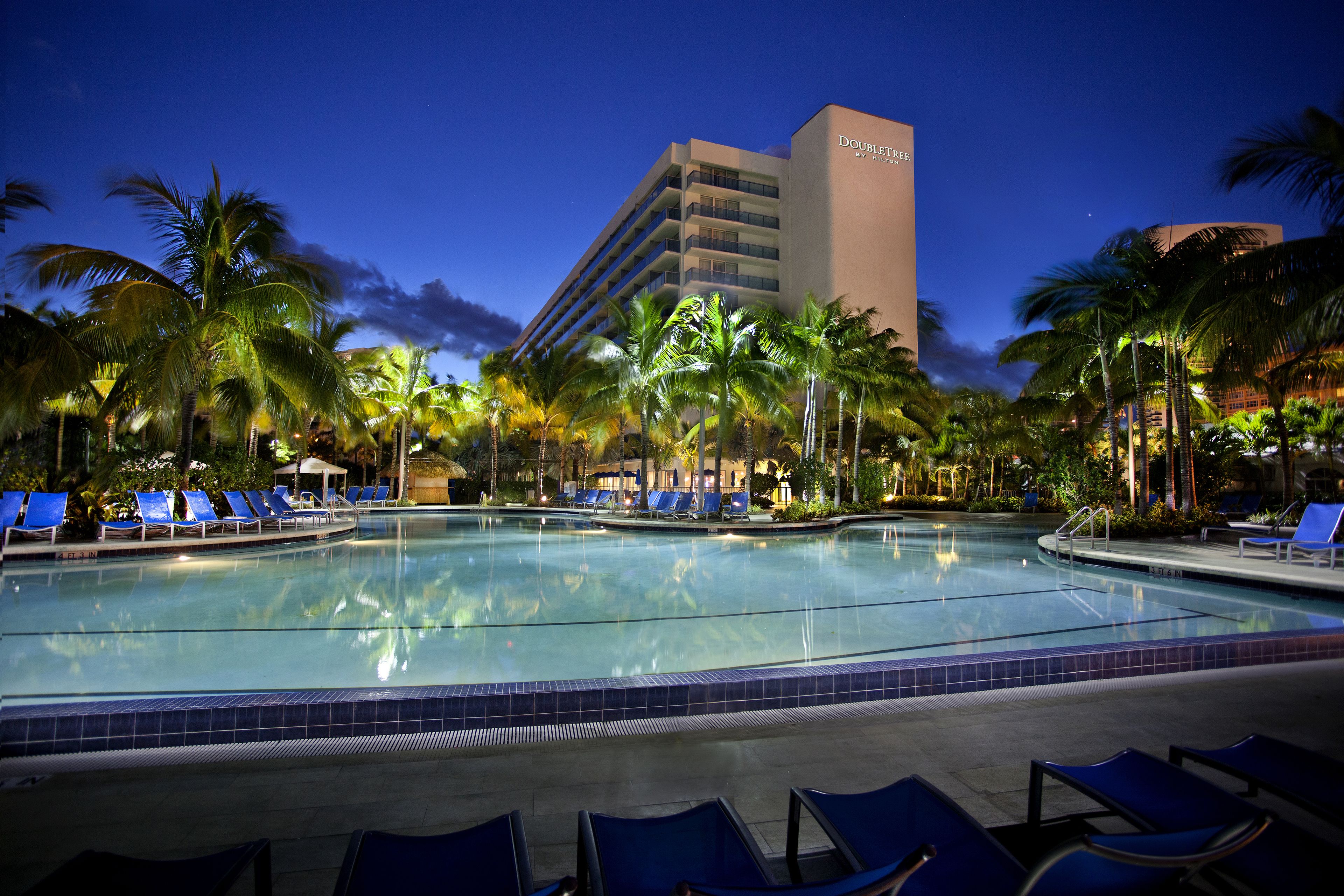 A hotel pool with blue lounge chairs, palm trees, and a building in the background.