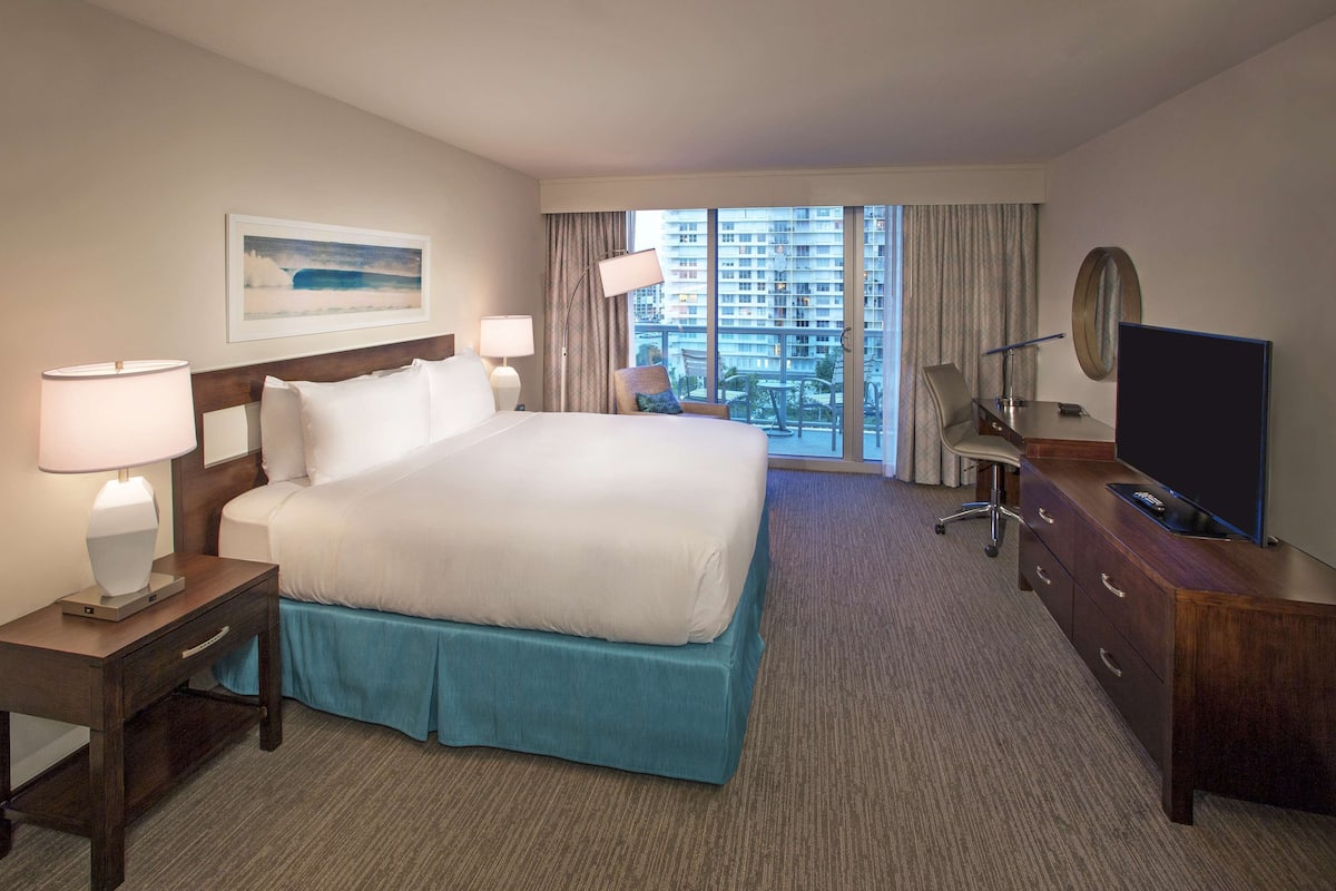 Ein Hotelzimmer mit einem großen Bett, einem Schreibtisch mit Fernseher, einem Sessel und Blick auf Gebäude durch das Fenster.
