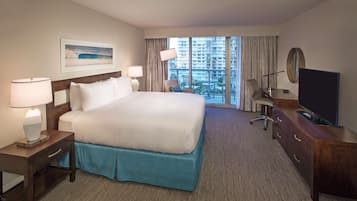 Quarto de hotel com uma cama grande, uma mesa de escritório com televisão, uma cadeira e vista para edifícios pela janela.