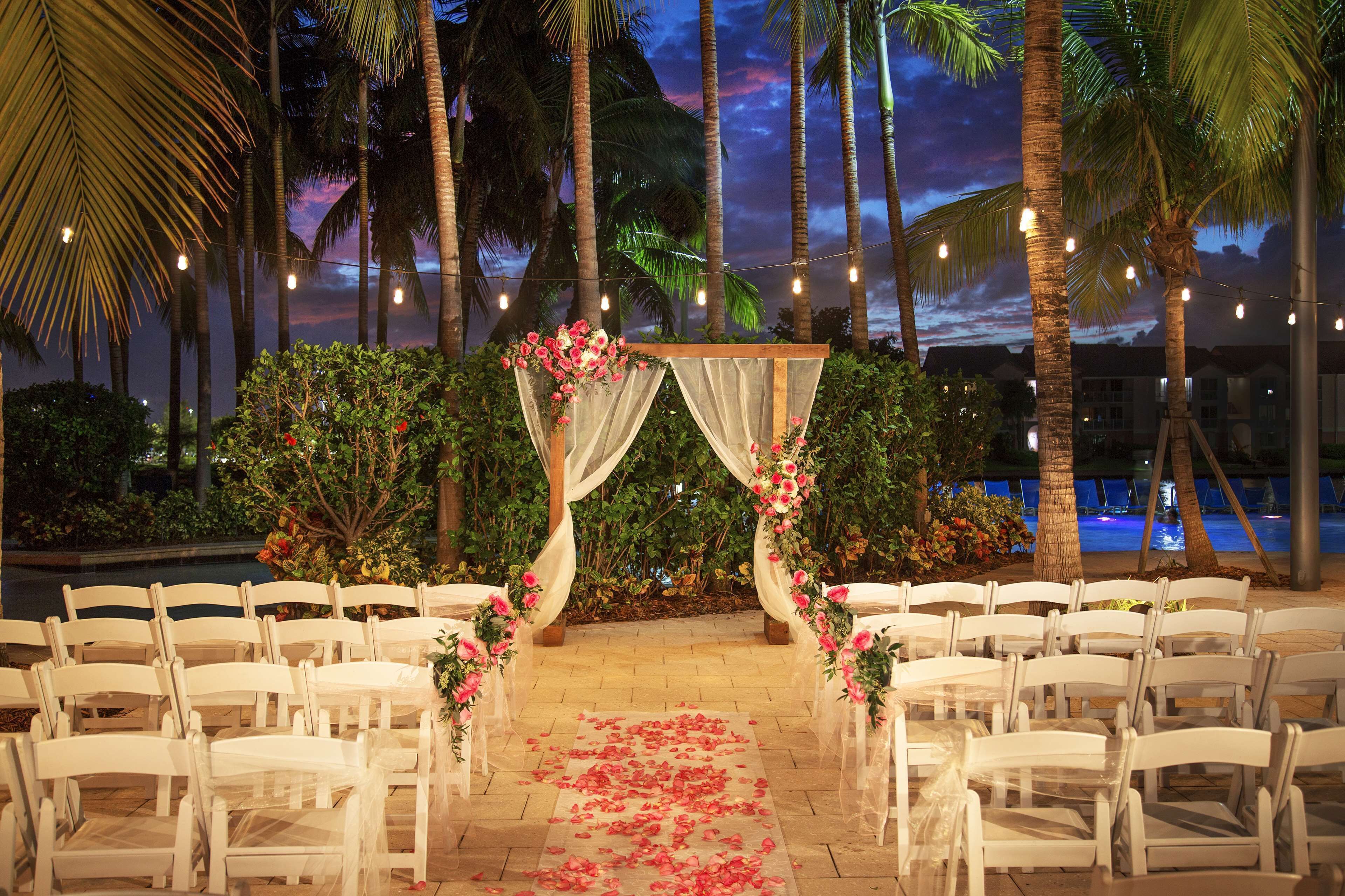 Une cérémonie de mariage en plein air, avec des chaises blanches disposées en rangées, une arche décorée de fleurs et, en arrière-plan, des palmiers et un ciel crépusculaire.