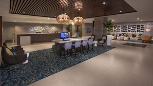 Un moderno vestíbulo de hotel con mostrador de recepción, zona de bar, asientos y un tapete azul estampado, además de un diseño de techo de madera.