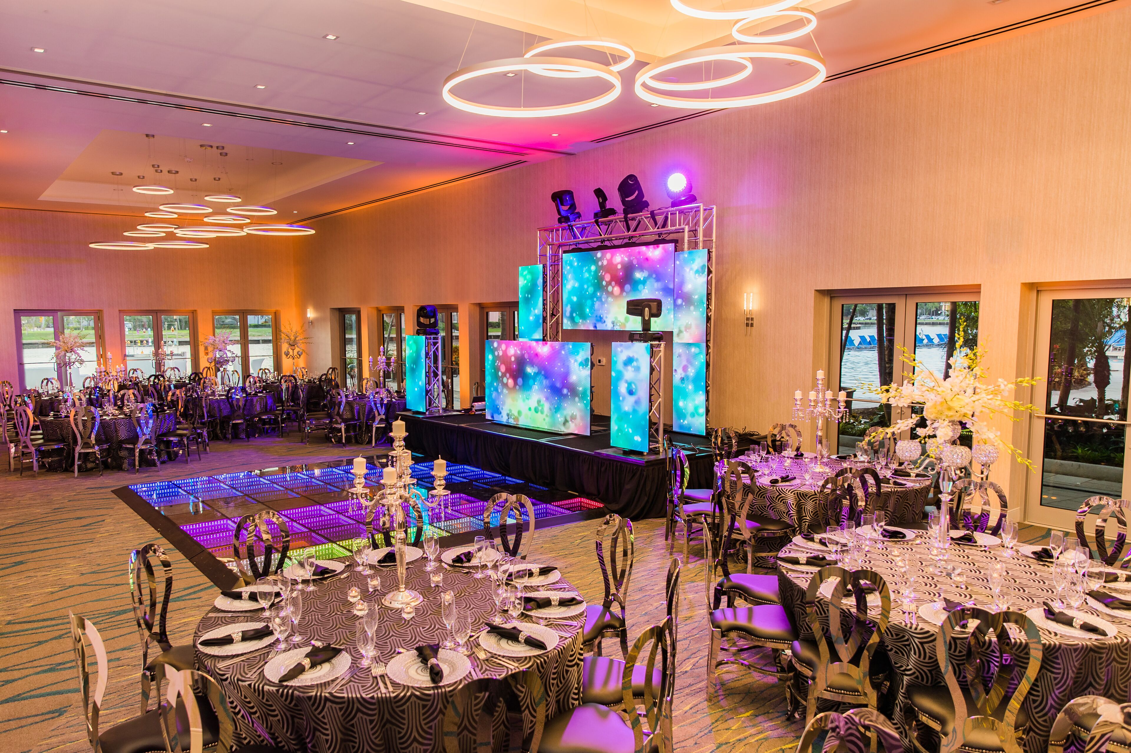 A banquet hall with round tables set for dining, a stage with a large screen, and a dance floor with LED lighting.