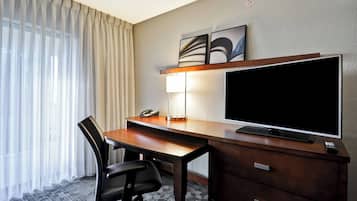 A hotel room with a wooden desk, a black office chair, a flat-screen TV, a lamp, and a telephone.