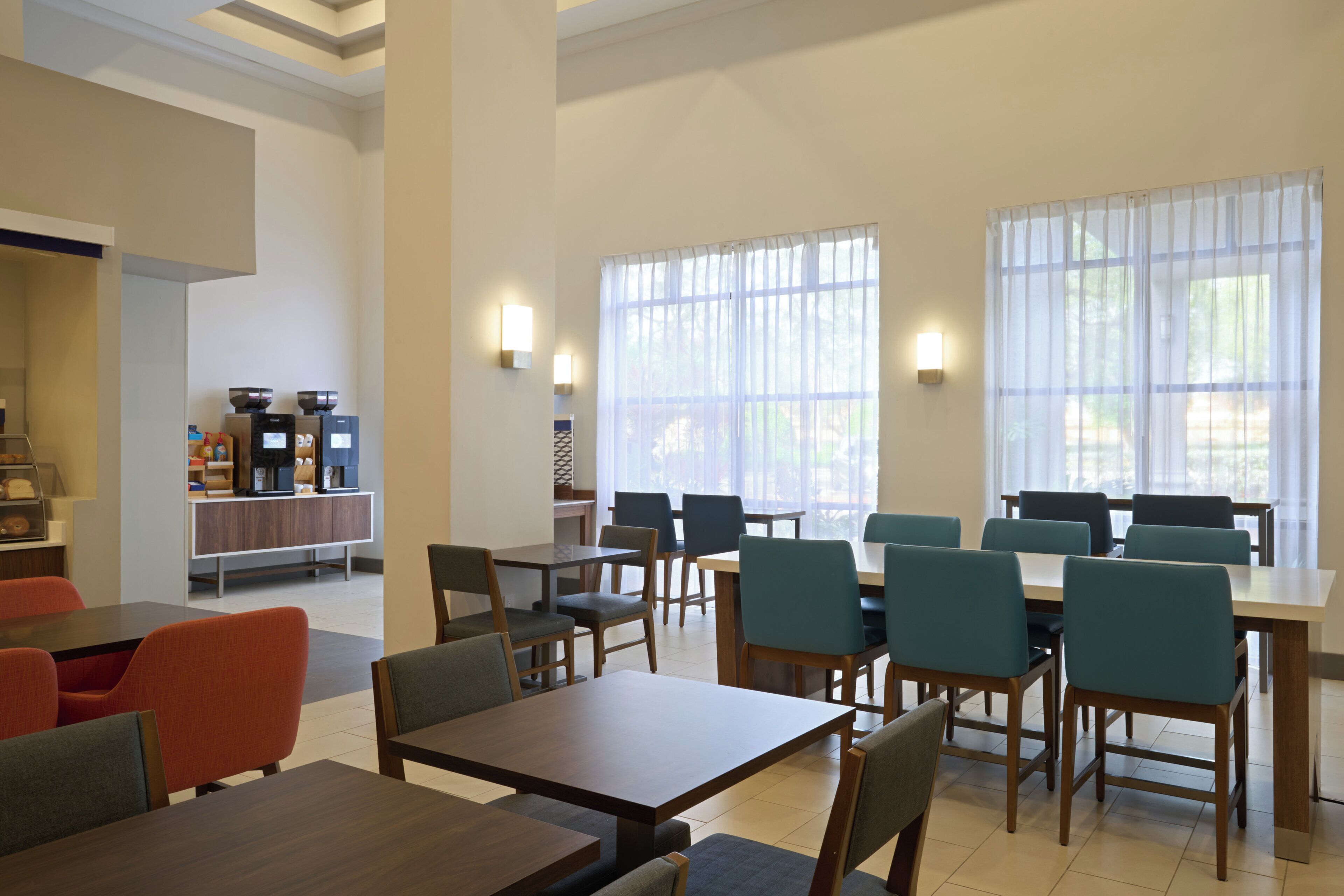 A dining area with tables and chairs, a counter with a coffee machine, and large windows with sheer curtains.