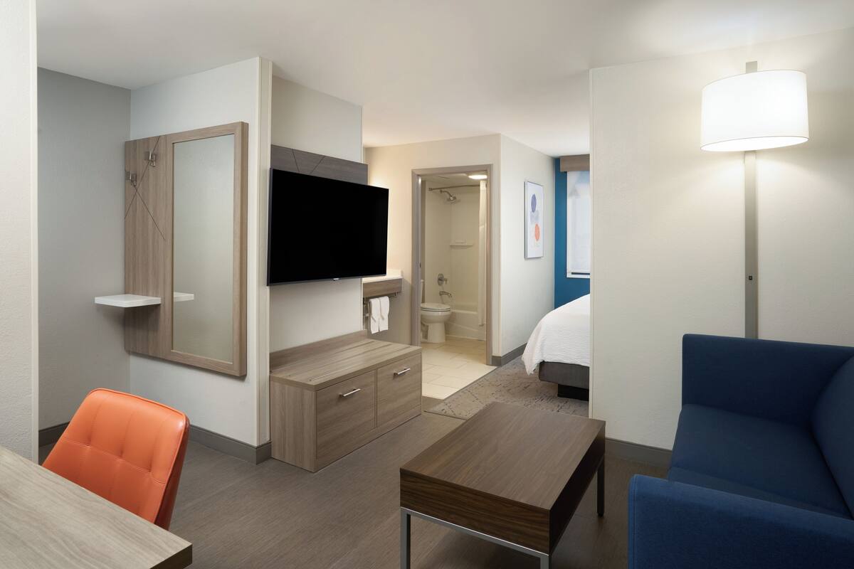 Ein modernes Hotelzimmer mit Flachbildfernseher, einem Holzhocker, einem blauen Sessel und einem Nachttisch.