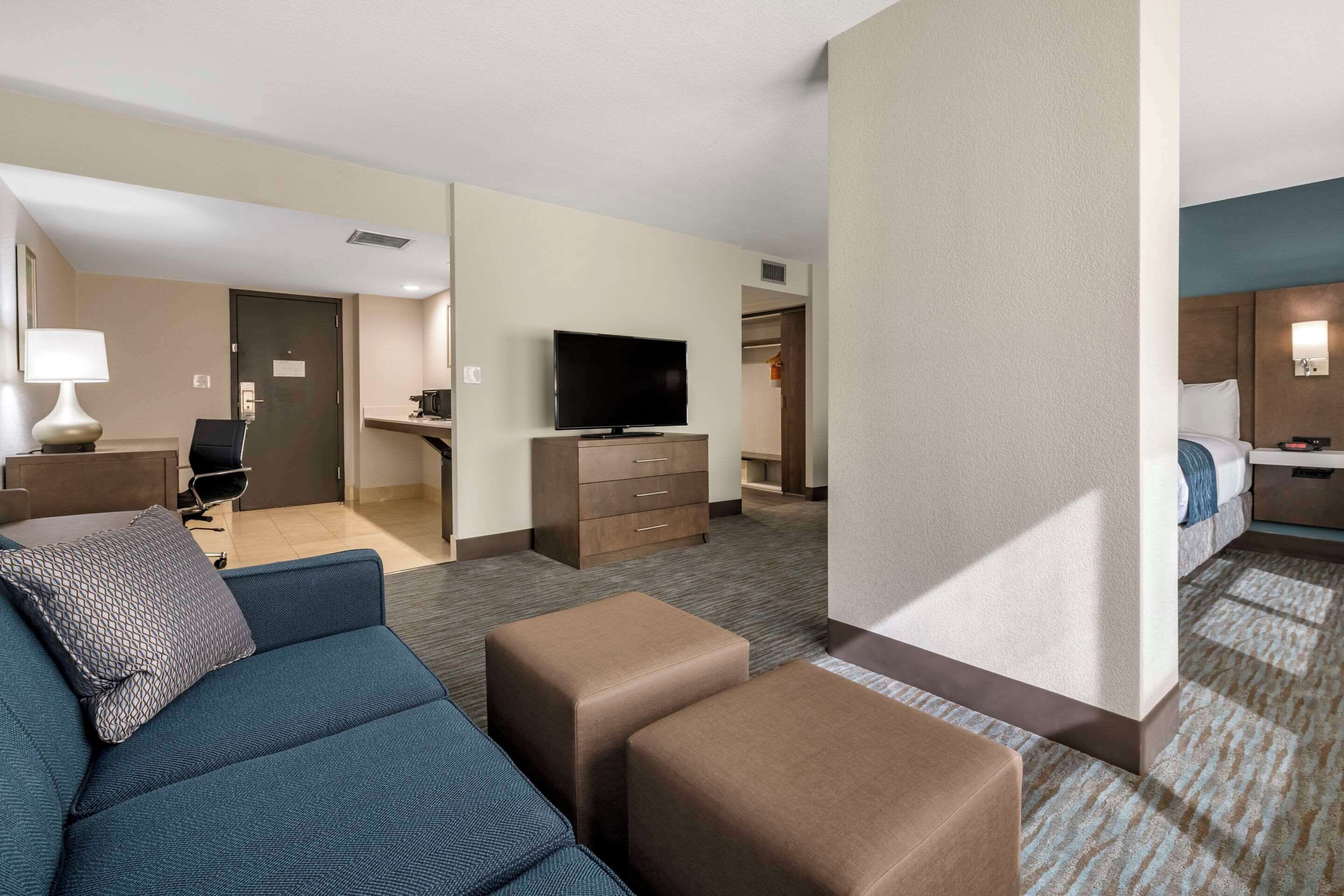 Ein Hotelzimmer mit Sofa, Hocker, Fernseher, Schreibtisch und Kleiderschrank.