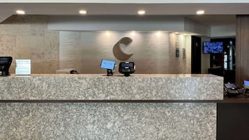 A hotel reception desk with a marble counter, a digital welcome screen, a check-in kiosk, and a television displaying a program.