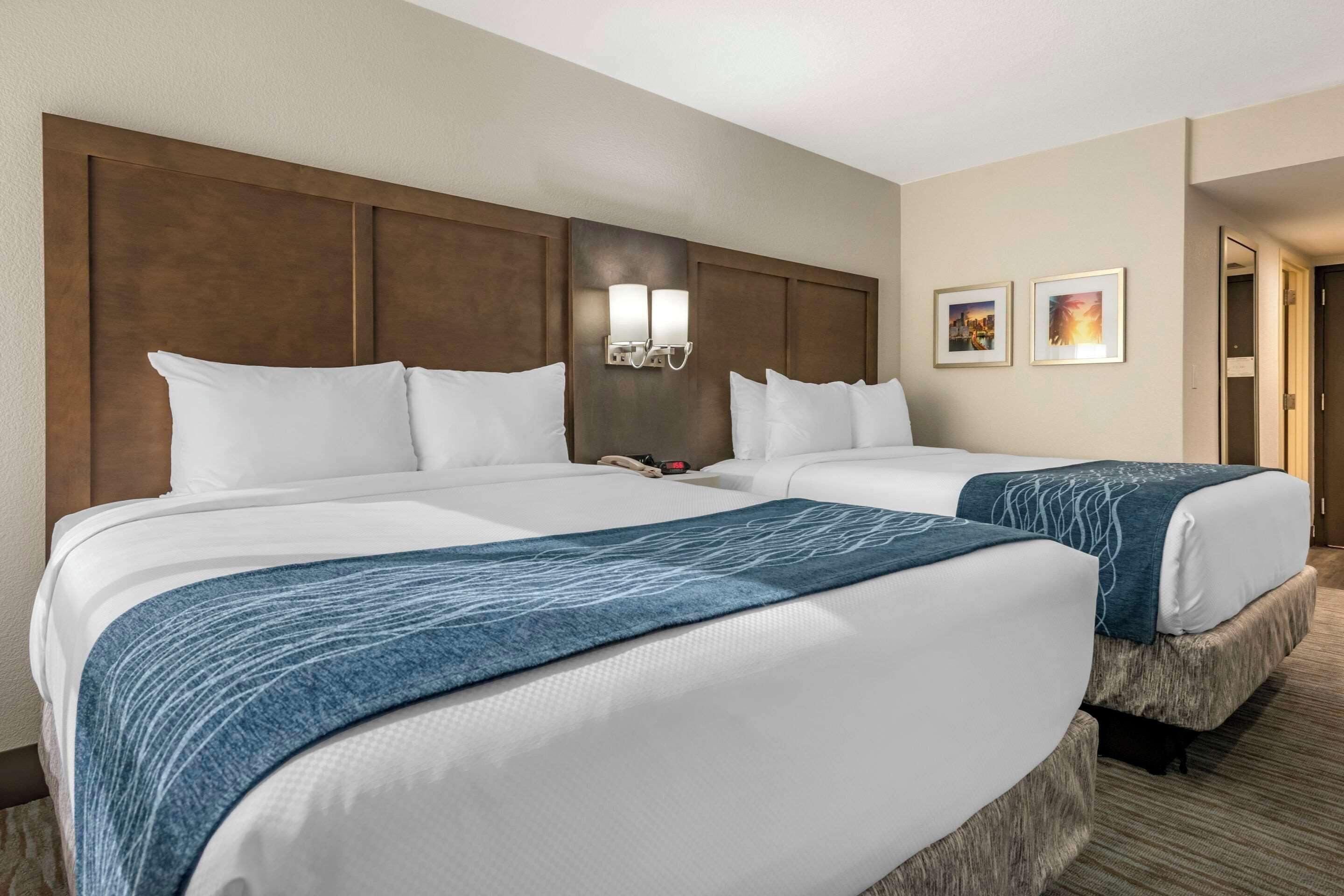 A hotel room with two beds, a headboard, wall-mounted lights, and two framed pictures.