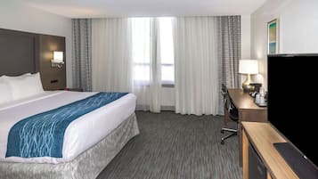 Quarto de hotel com uma cama grande, uma escrivaninha com televisão e uma janela com cortinas.