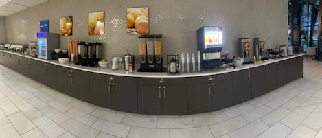 Una moderna zona de recepción de hotel con un mostrador curvo, máquinas de café y una estación de bebidas de autoservicio.