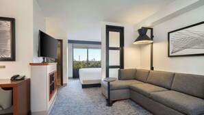 A modern hotel room with a large bed, a sofa, a desk with a phone, a TV, and a large window offering a city view. - Hyatt Place Busch Gardens (Tampa)