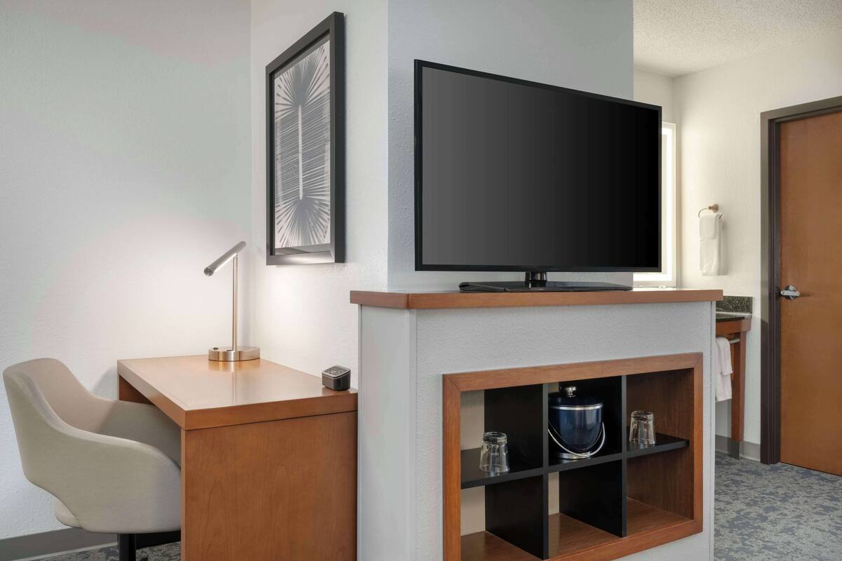 A hotel room with a wooden desk, a chair, a flat-screen TV mounted above the desk, a framed picture on the wall, and a small shelf with a kettle and other items.