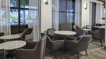 A hotel lobby with round tables and chairs, large windows with curtains, and a carpeted floor.
