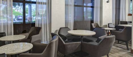 A hotel lobby with round tables and chairs, large windows with curtains, and a carpeted floor.