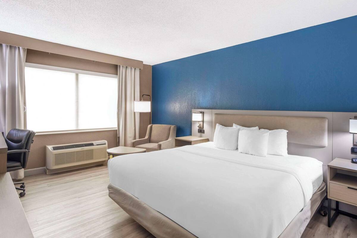 A hotel room with a large bed, a desk, a chair, a lamp, and a window.