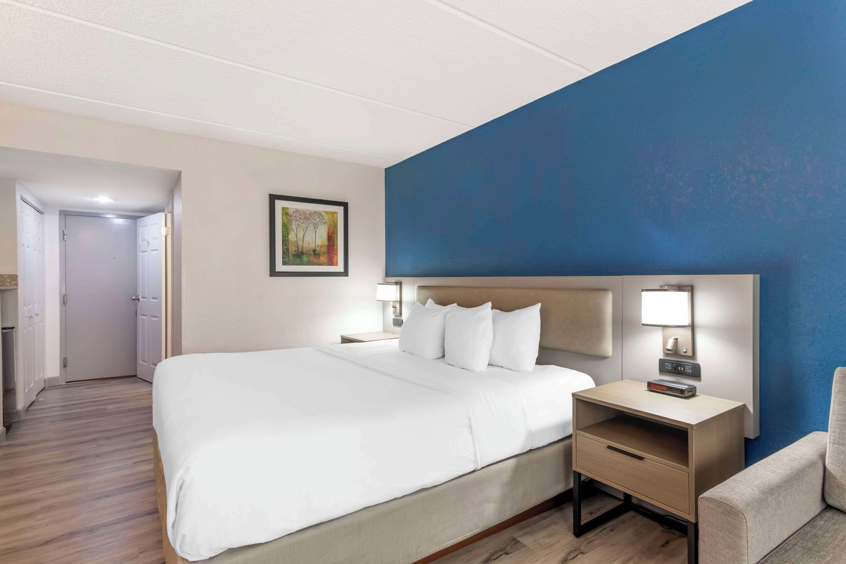 Una habitación de hotel con una cama grande, una mesita de noche con lámpara, una silla y un cuadro en la pared.