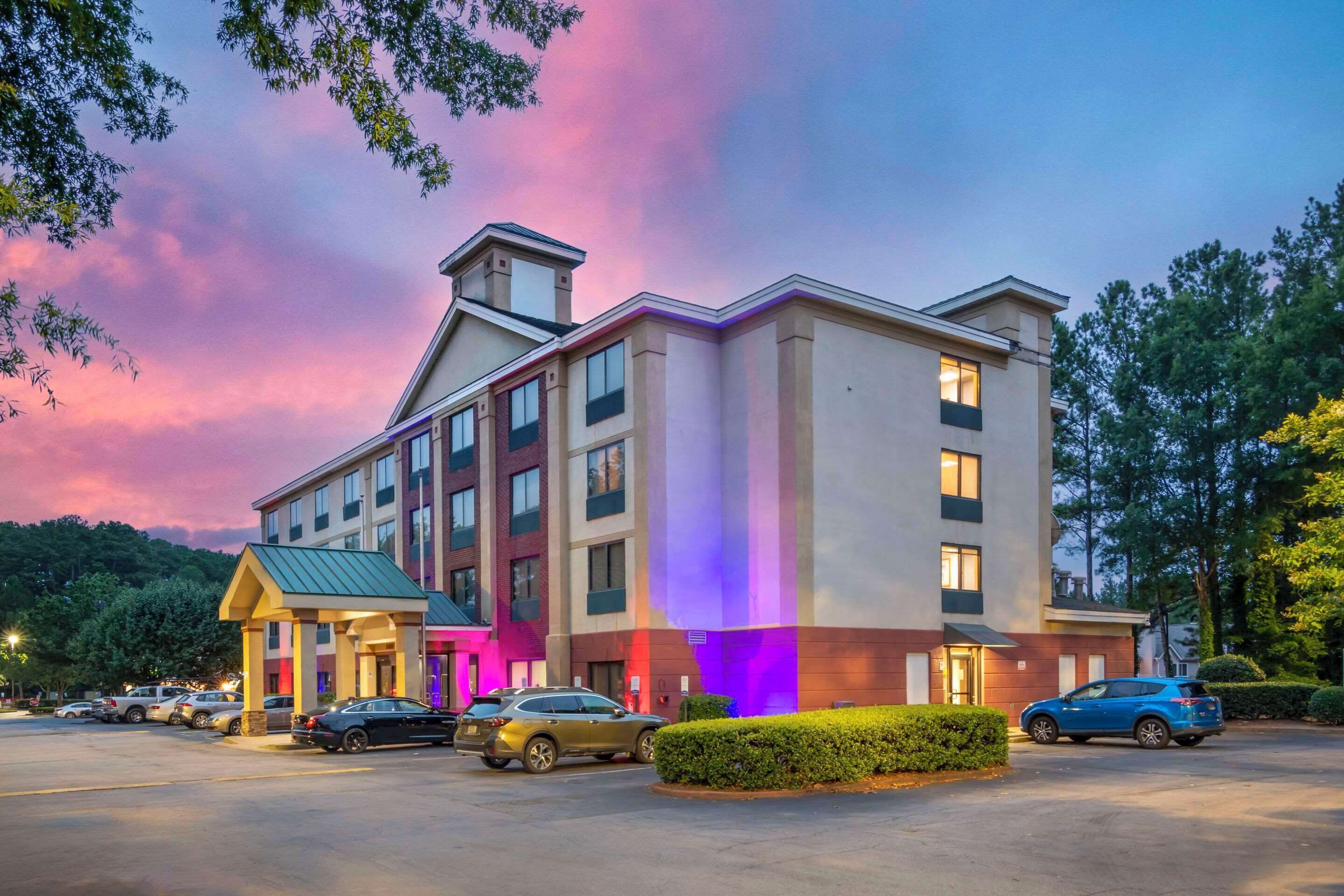 A multi-story hotel with a central entrance, parking lot, and surrounding greenery.