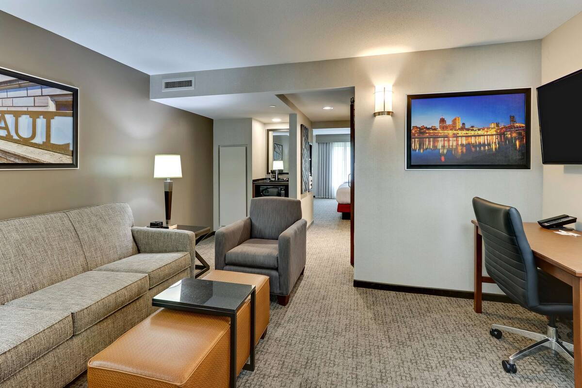 A hotel room with a sofa, armchair, desk, and a cityscape painting.