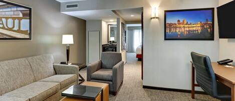 A hotel room with a sofa, armchair, desk, and a cityscape painting.