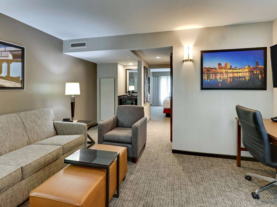 A hotel room with a sofa, armchair, desk, and a cityscape painting.