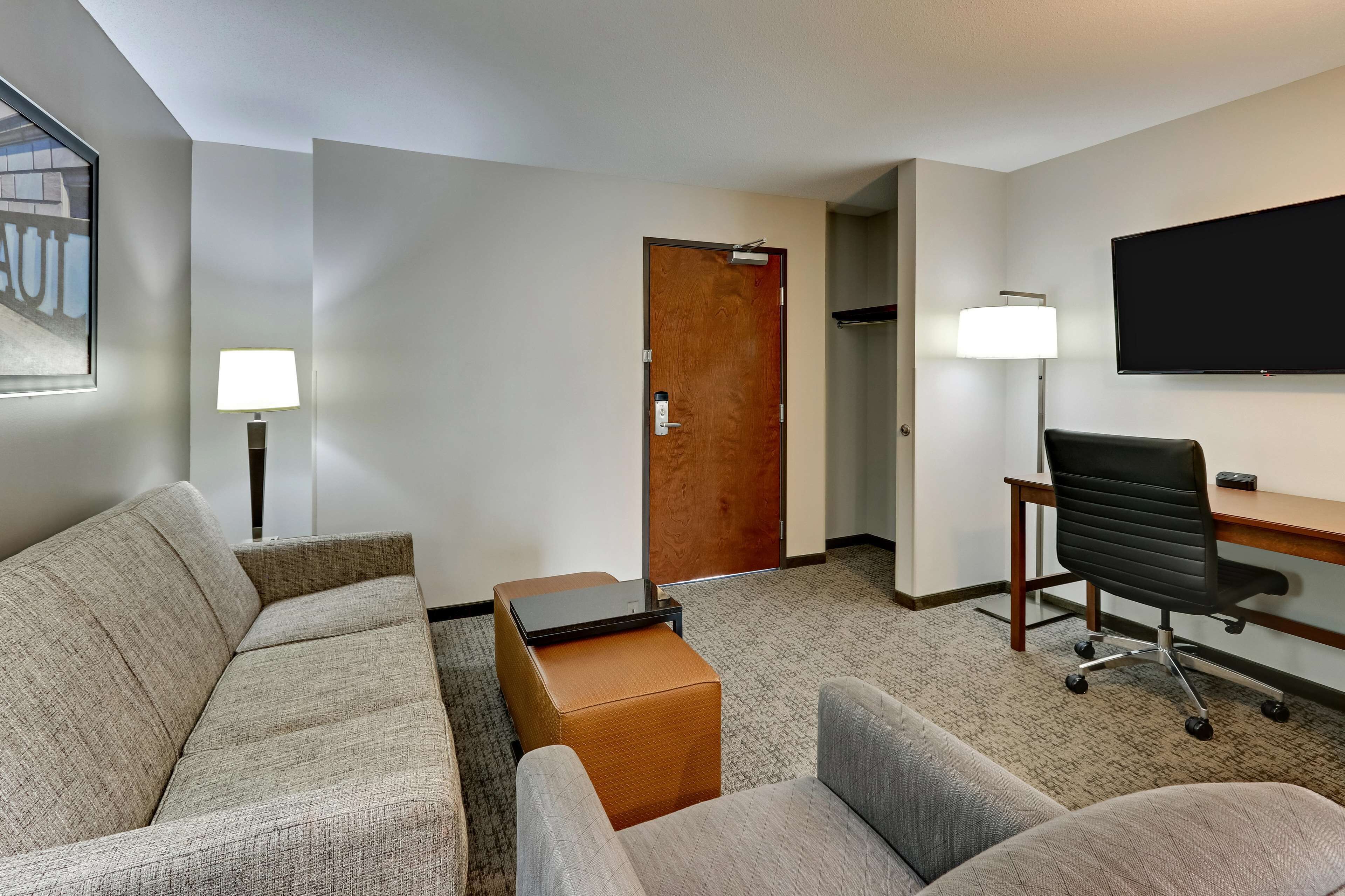 A hotel room with a sofa, a small table, a desk with a chair, a flat-screen TV, and a door.