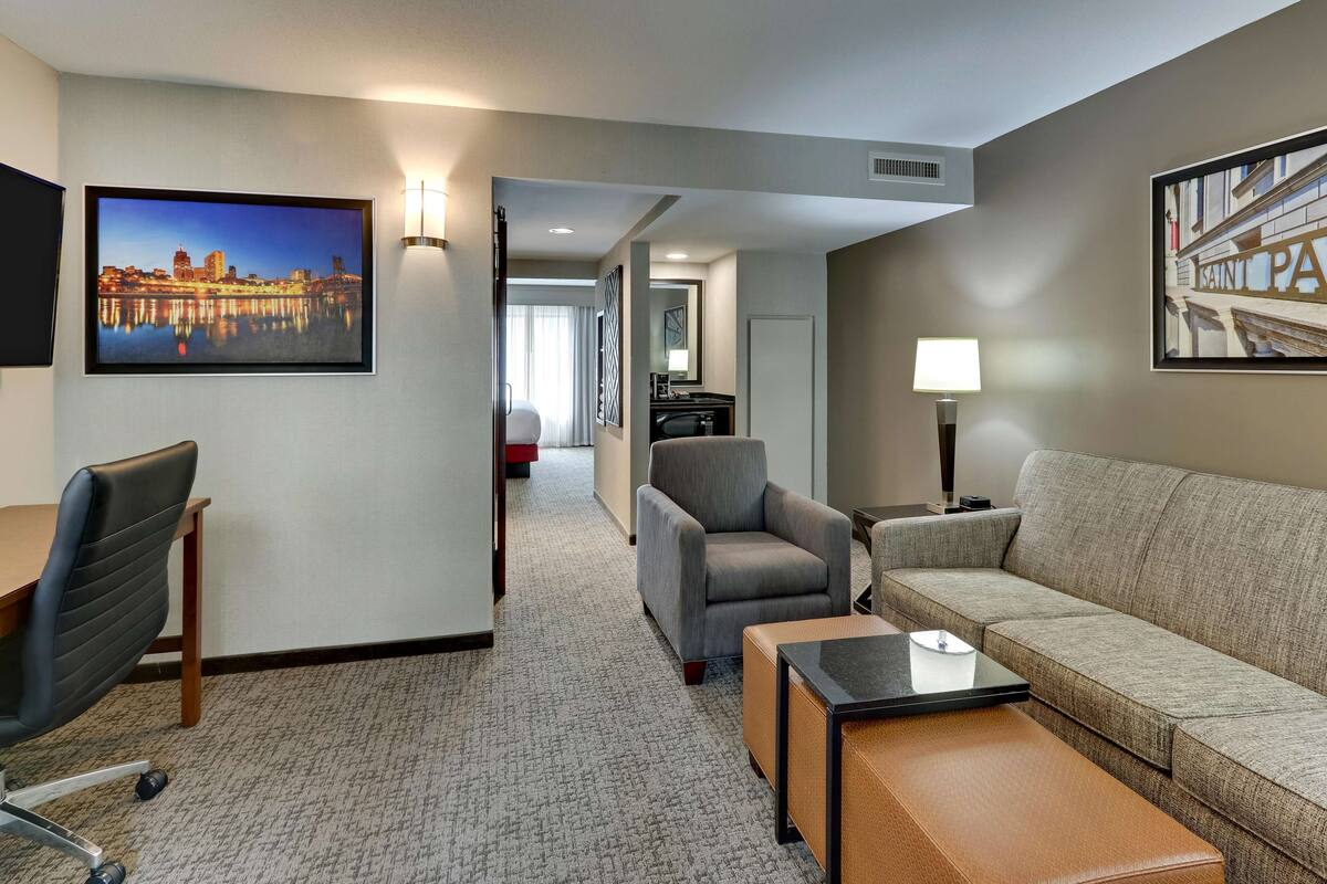A hotel room with a sofa, armchair, desk, and a cityscape painting.
