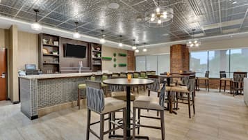 Um restaurante moderno com teto alto, painéis metálicos no teto e luminárias pendentes. Possui diversas mesas e cadeiras para refeições, um bar e uma televisão na parede.