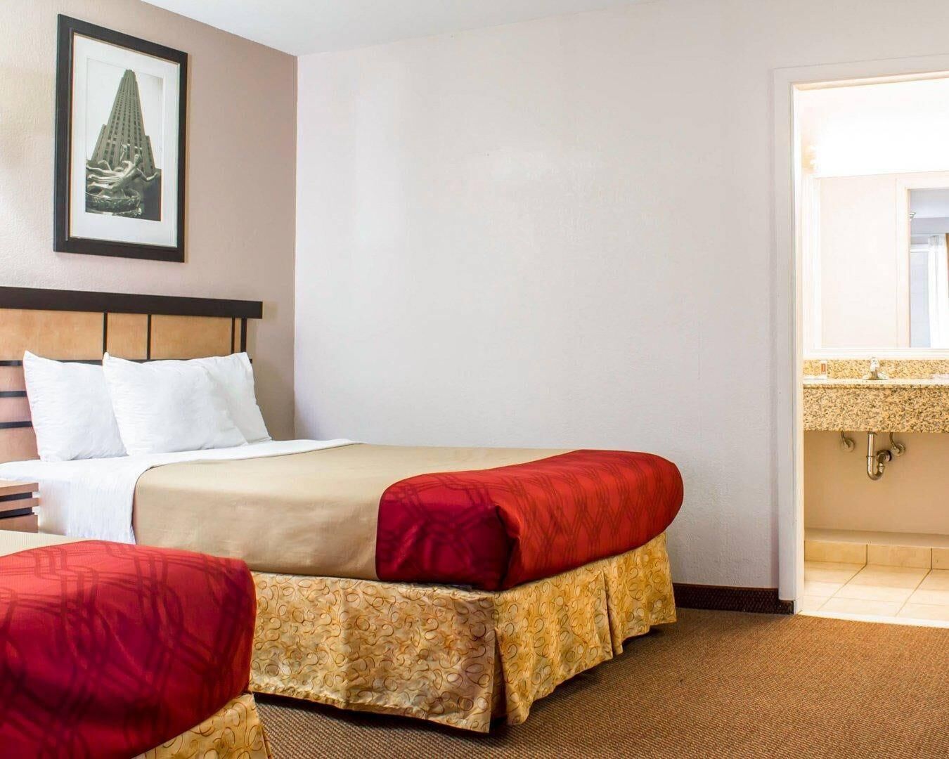 A hotel room with a bed, a headboard, a framed picture on the wall, and a bathroom visible through an open door.